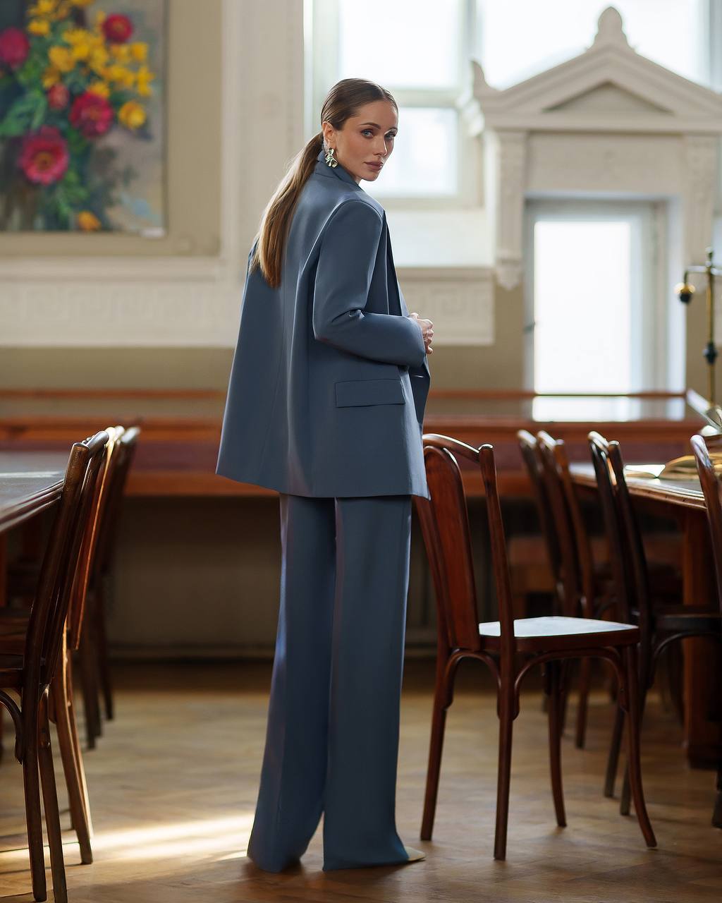 Woman in a blue suit standing in a room with wooden chairs and tables.