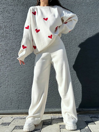 Person wearing a white sweater with red heart patterns and white pants against a gray wall.
