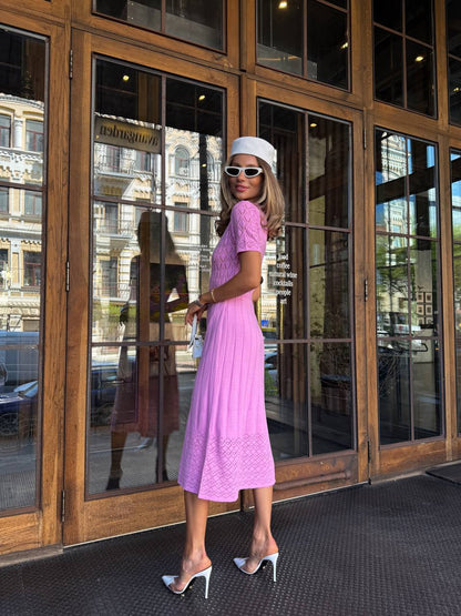 Woman in a pink dress and white hat standing in front of large glass doors.