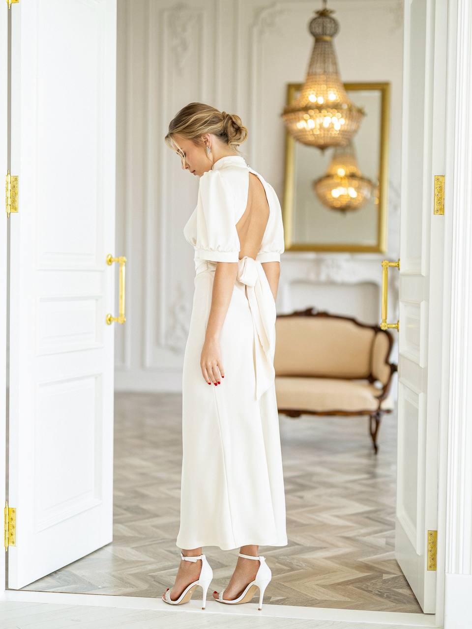 A woman wearing a white midi dress with puff sleeves and a side tie detail, standing in an elegant room framed by open white doors.