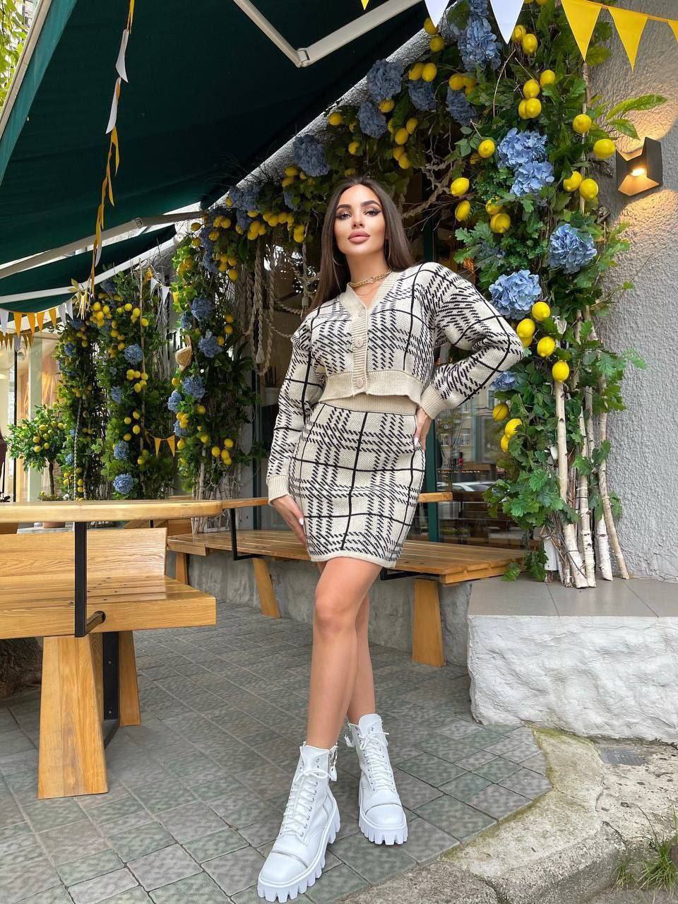 Woman in a plaid outfit standing in front of a decorated outdoor setting with flowers and greenery.