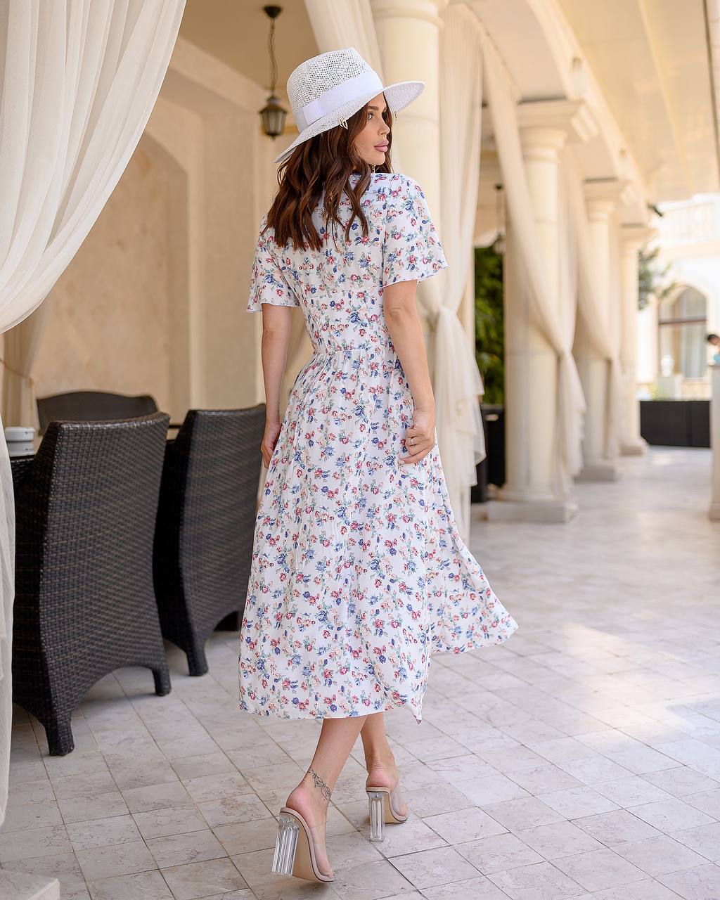 Woman in a floral dress and white hat standing in an outdoor setting with columns and tables.