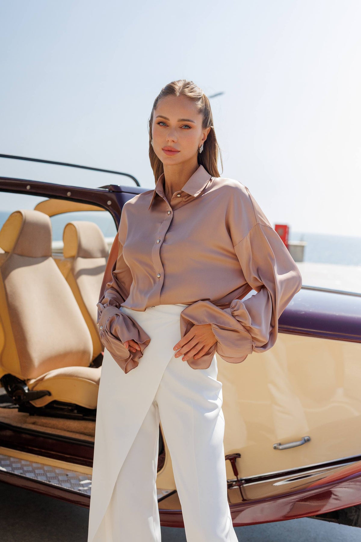 Woman in a beige blouse and white pants standing next to a vintage car.