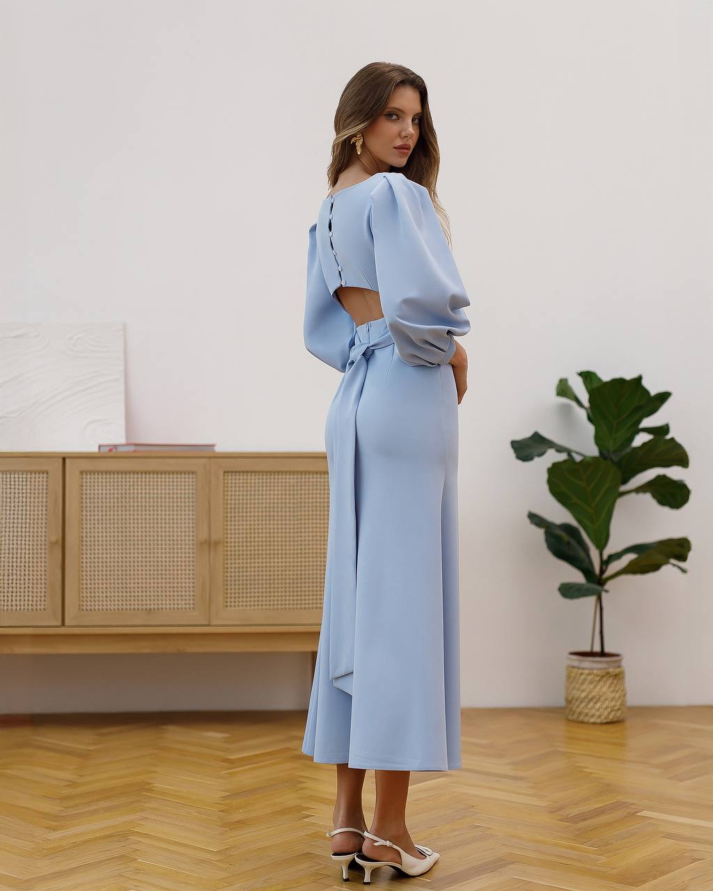 Woman wearing a light blue dress standing in a room with wooden flooring and a plant.
