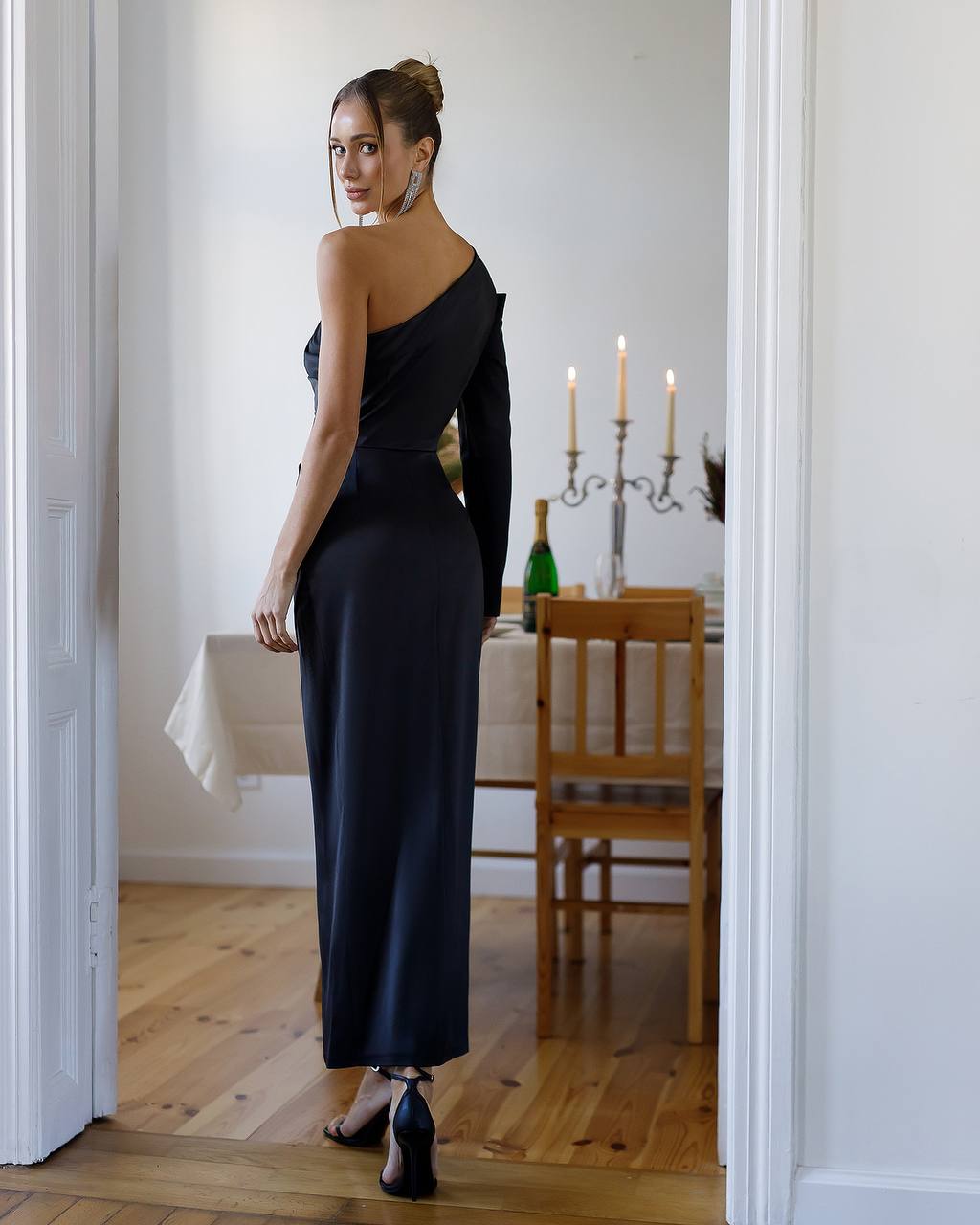Woman in a black one-shoulder dress standing in a room with a dining table and candles.