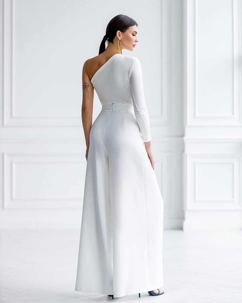 Woman wearing a white one-shoulder jumpsuit against a white wall.