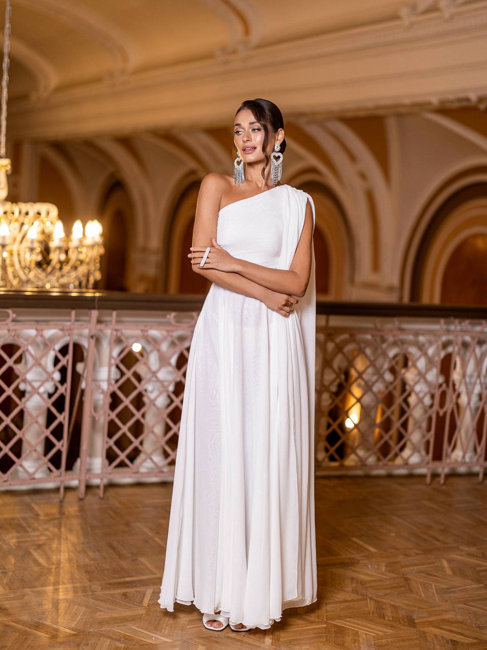 Woman in a white one-shoulder gown standing in an elegant indoor setting with chandeliers.