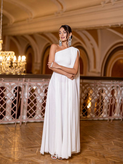 Woman in a white one-shoulder gown standing in an elegant indoor setting with chandeliers.