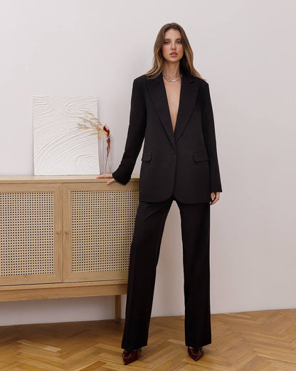 Woman wearing a black suit standing in a room with wooden flooring and a light-colored wall.