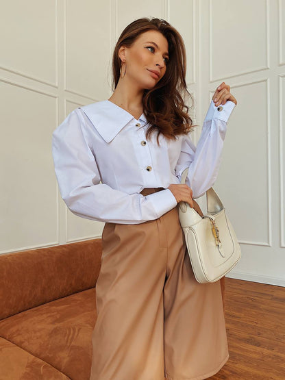 Woman in a white blouse and beige pants holding a cream handbag against a neutral wall.