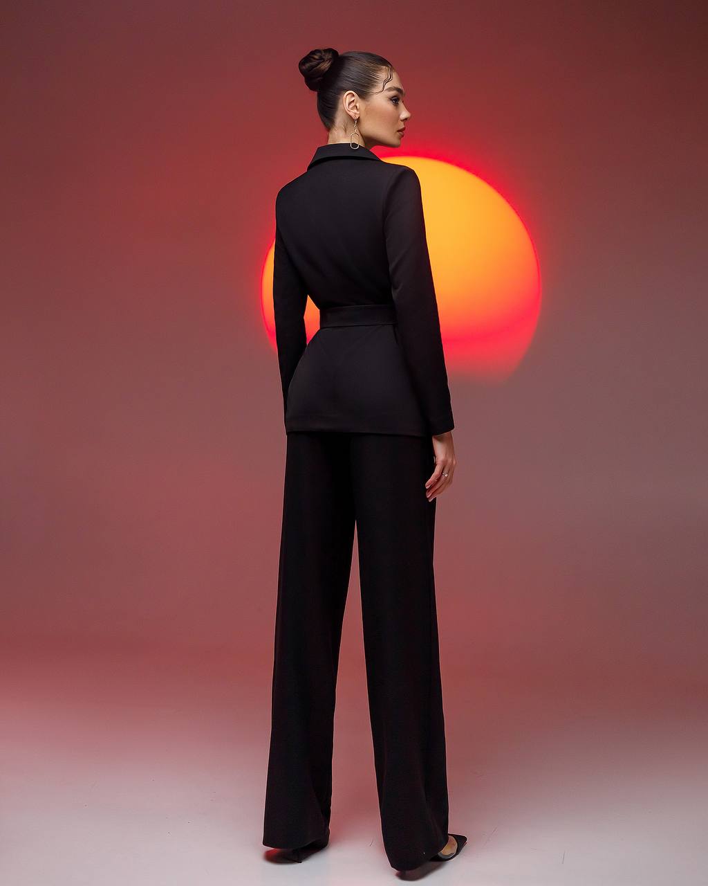Woman in a black suit standing with her back to the camera against a red and orange sunset background.