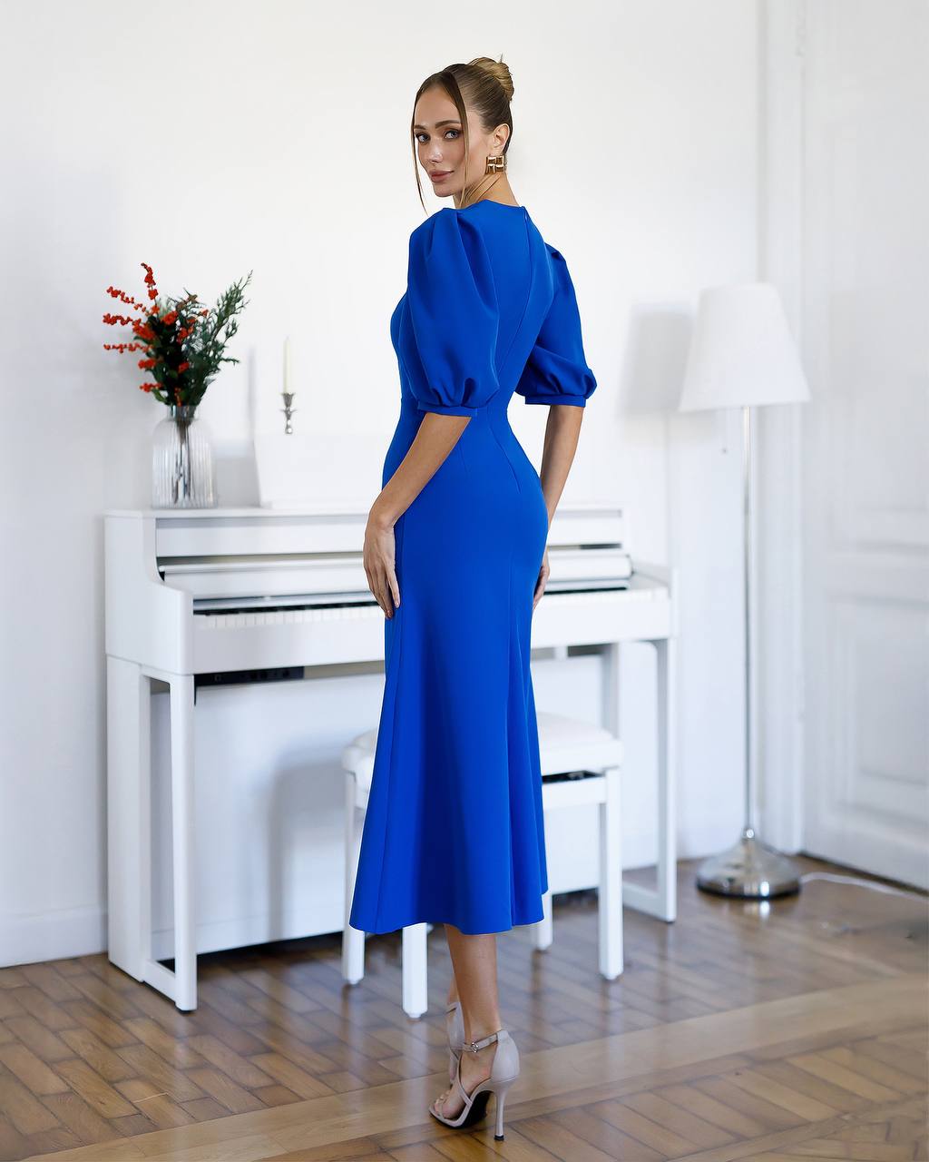 Woman in a blue dress standing in a room with a white piano and lamp.