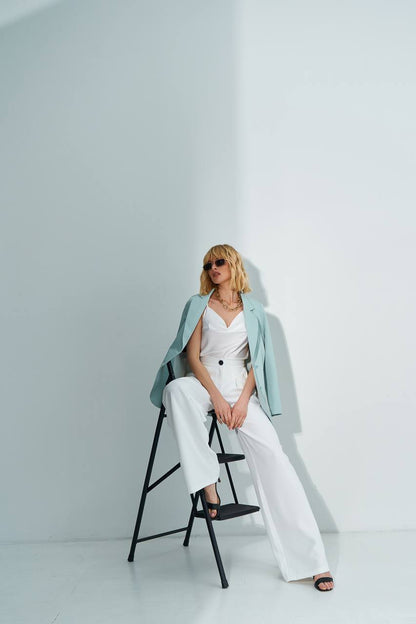 Woman in a white outfit with a light blue jacket sitting on a black stool against a white background