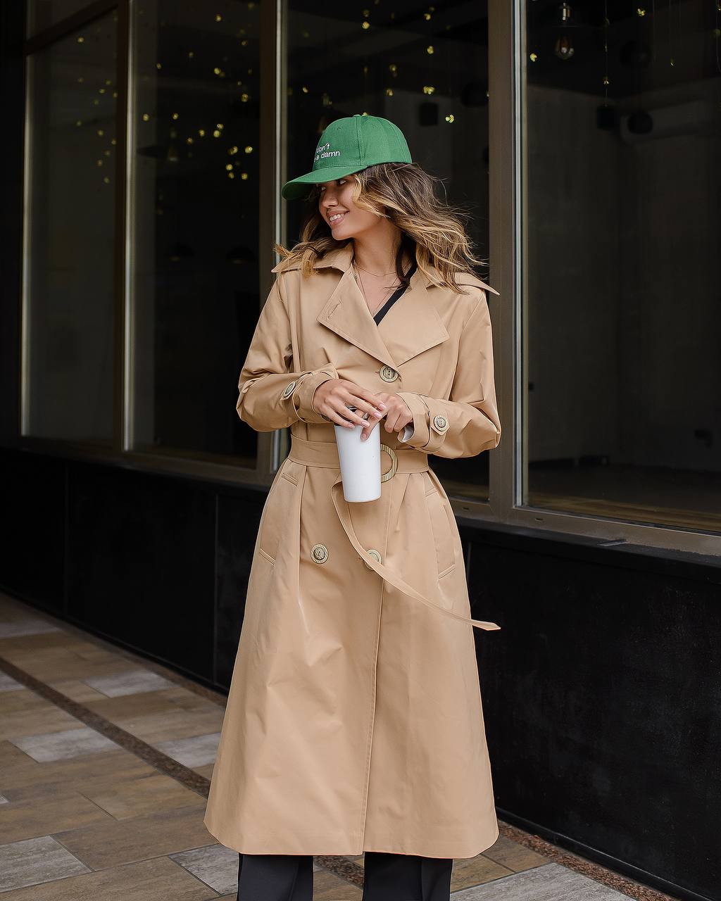 Woman in a beige trench coat and green cap holding a white cup, standing in front of a dark glass building.