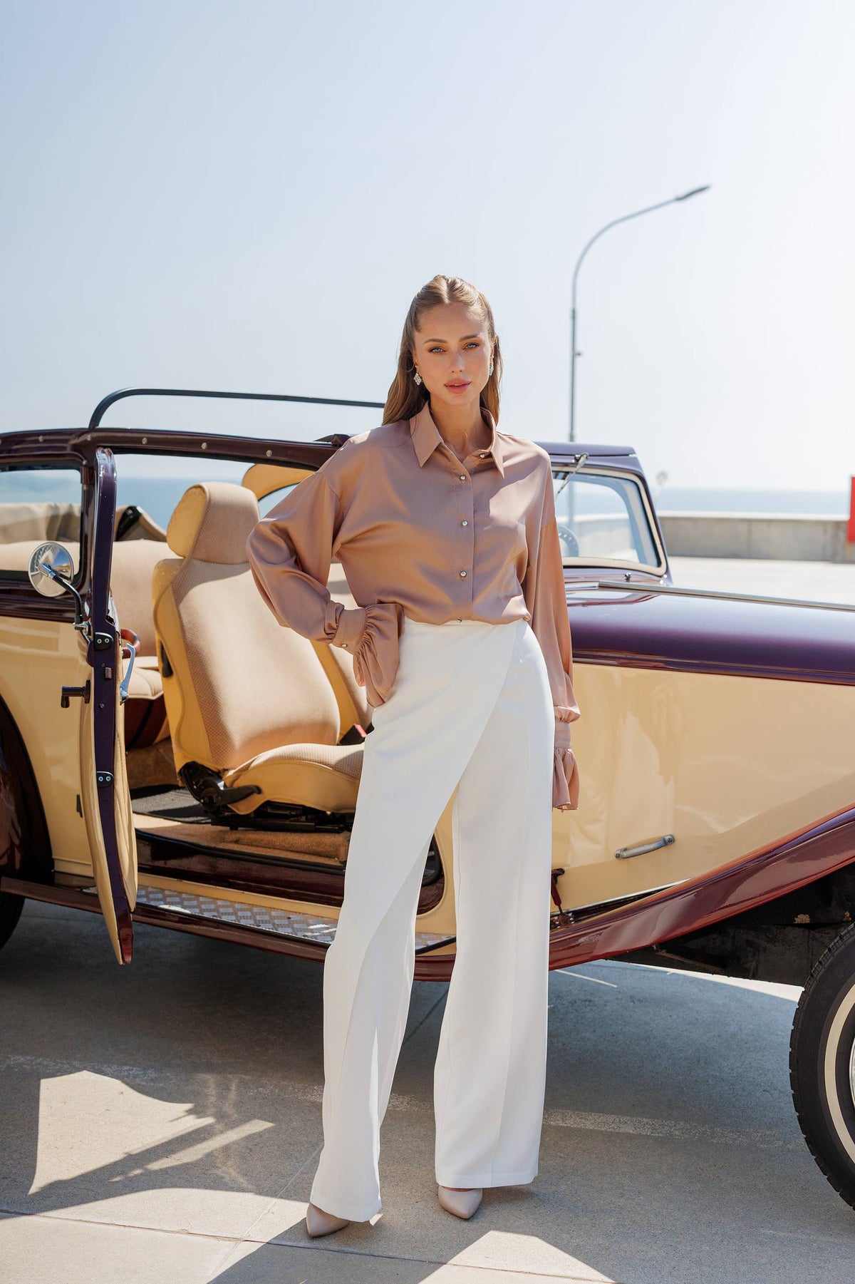 Woman standing next to a vintage car with an open door, wearing a light-colored blouse and white pants.