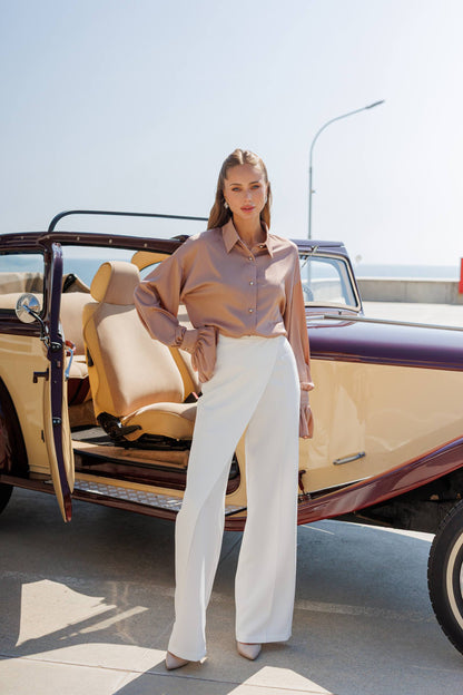 Woman standing next to a vintage car with an open door, wearing a light-colored blouse and white pants.