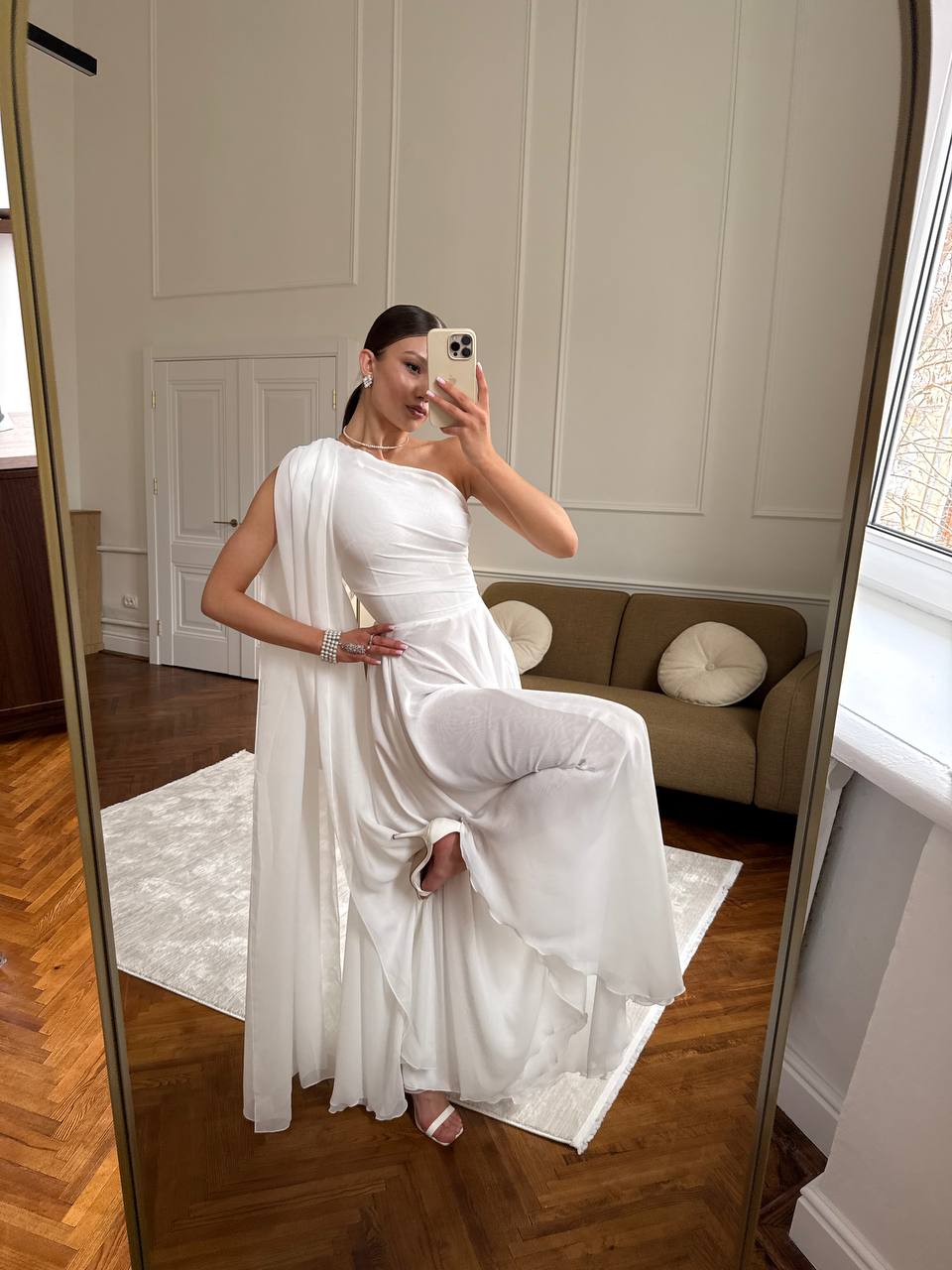 Woman in a white dress taking a mirror selfie in a room with a couch and window.