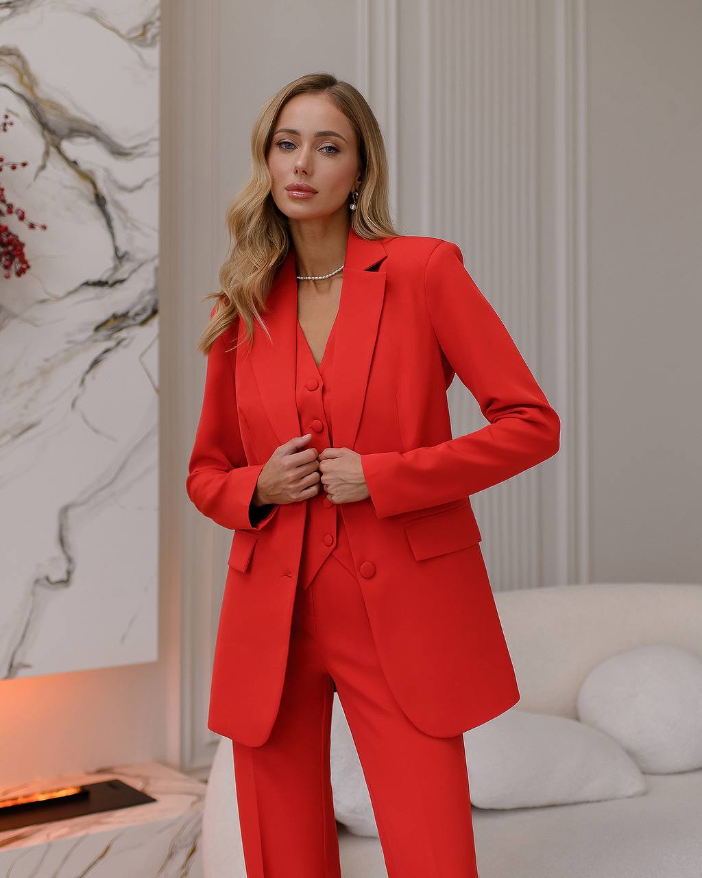 Woman wearing a red suit in an indoor setting with a marble wall and white sofa.