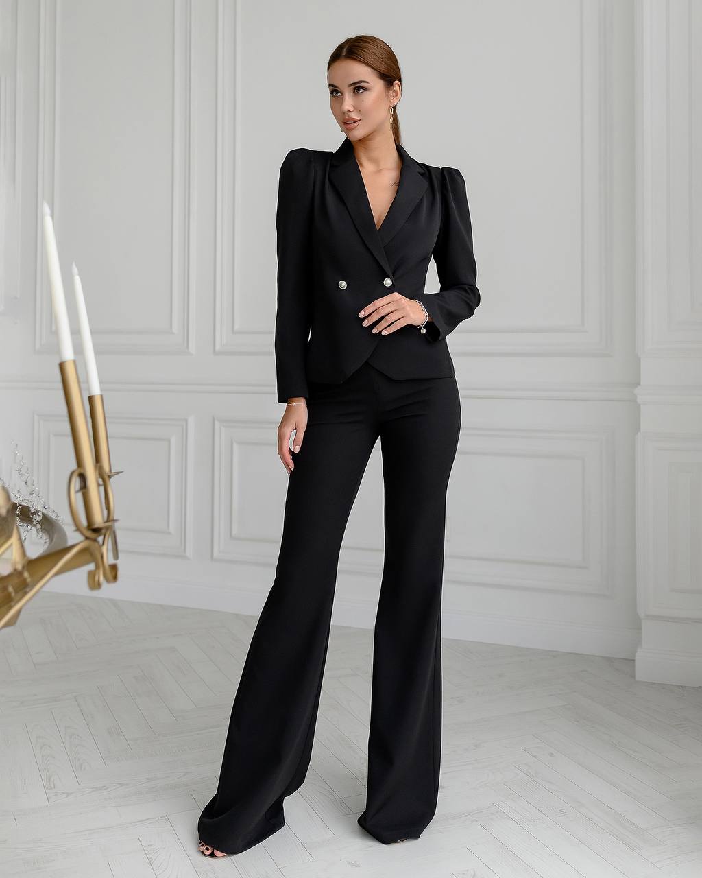 Woman wearing a black suit standing in a stylish room with white walls and a decorative wall piece.