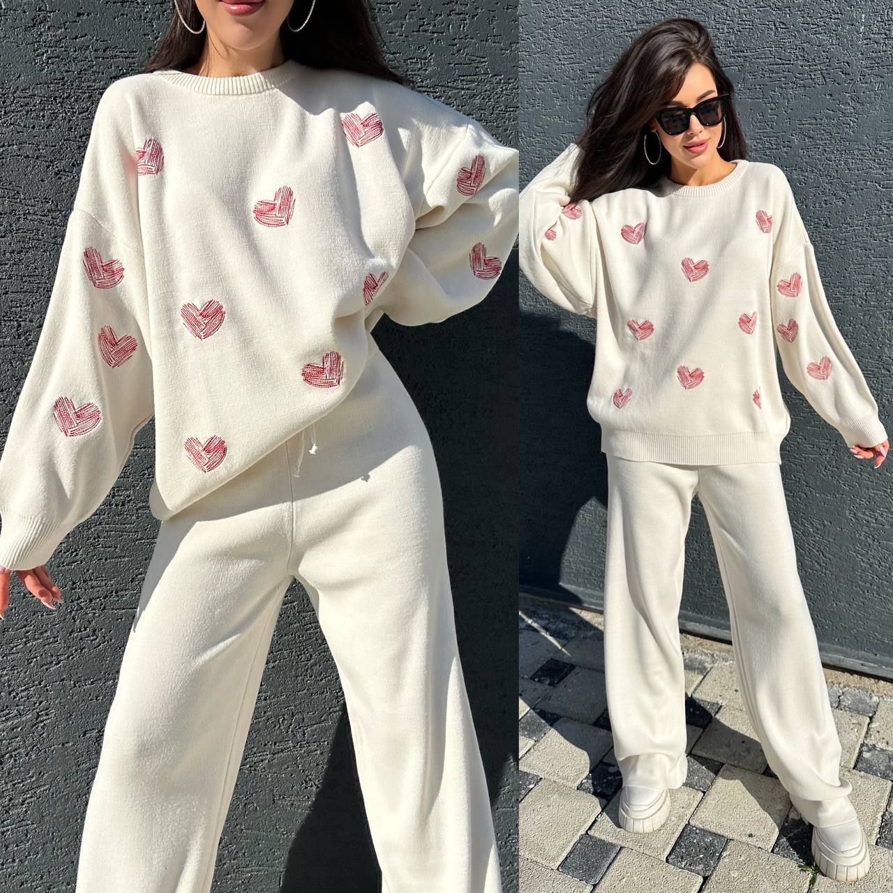 Woman wearing a white sweater with pink heart patterns and white pants against a gray wall.