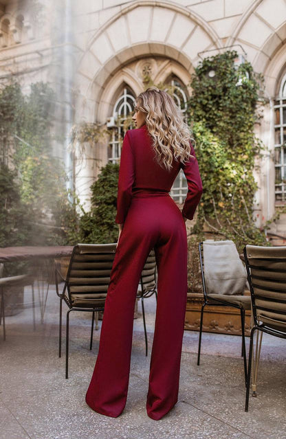 Person wearing a burgundy jumpsuit standing in an outdoor setting with architectural elements and greenery.
