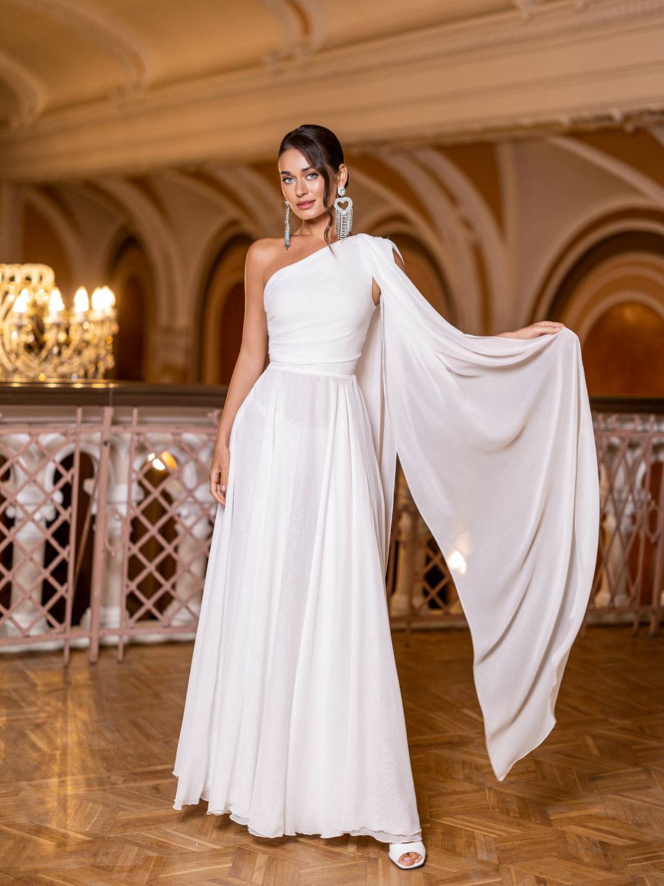 Woman in a white one-shoulder gown standing in an elegant indoor setting.