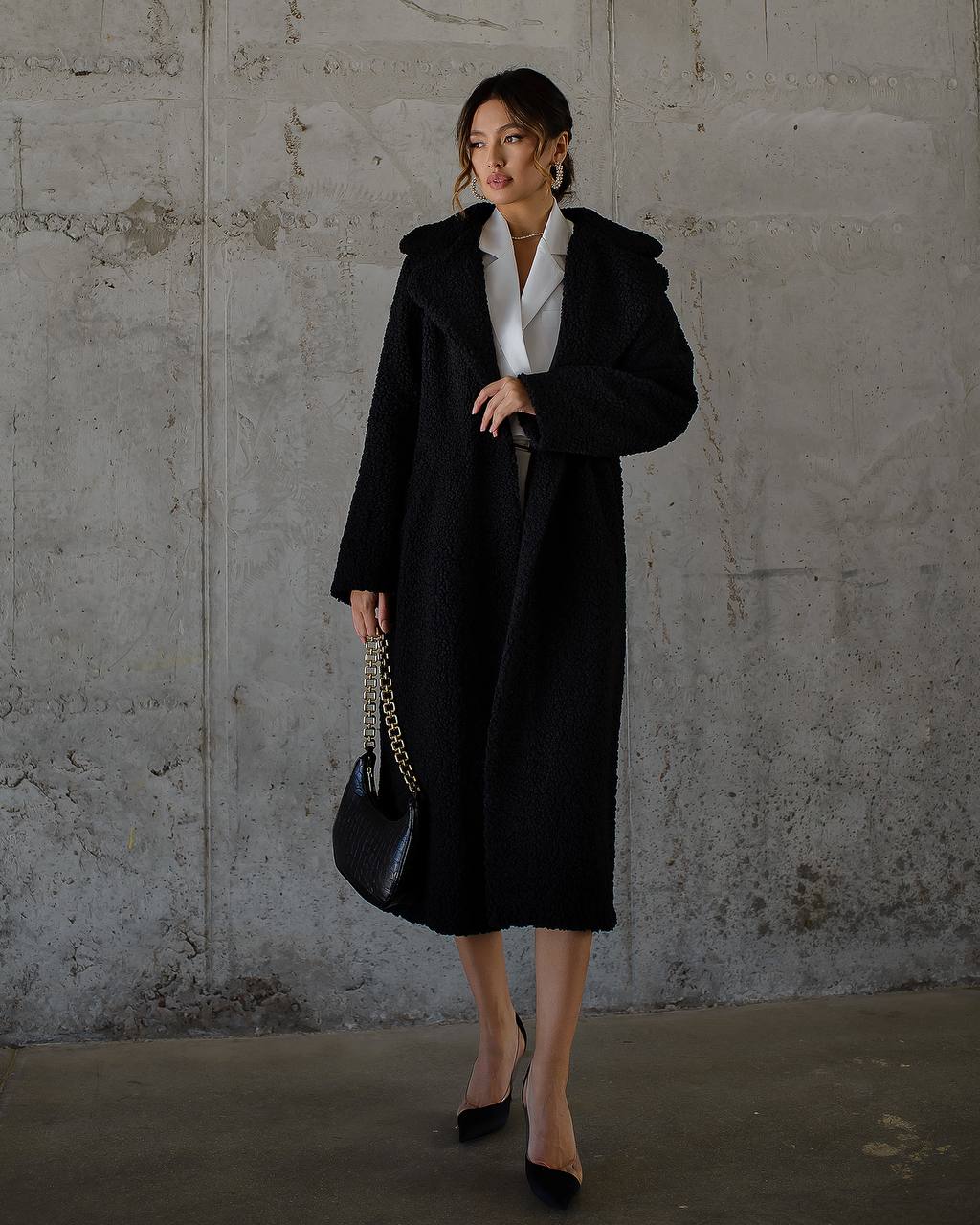 Woman wearing a black coat and holding a black handbag against a concrete wall.