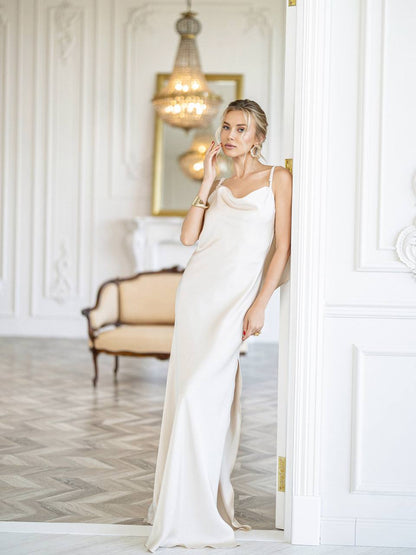 A woman wearing a floor-length cream satin gown with spaghetti straps and a cowl neckline, leaning against a white paneled door in an elegant room with a chandelier and upholstered bench.