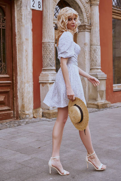 Woman in a white dress holding a straw hat, standing in front of an ornate building.