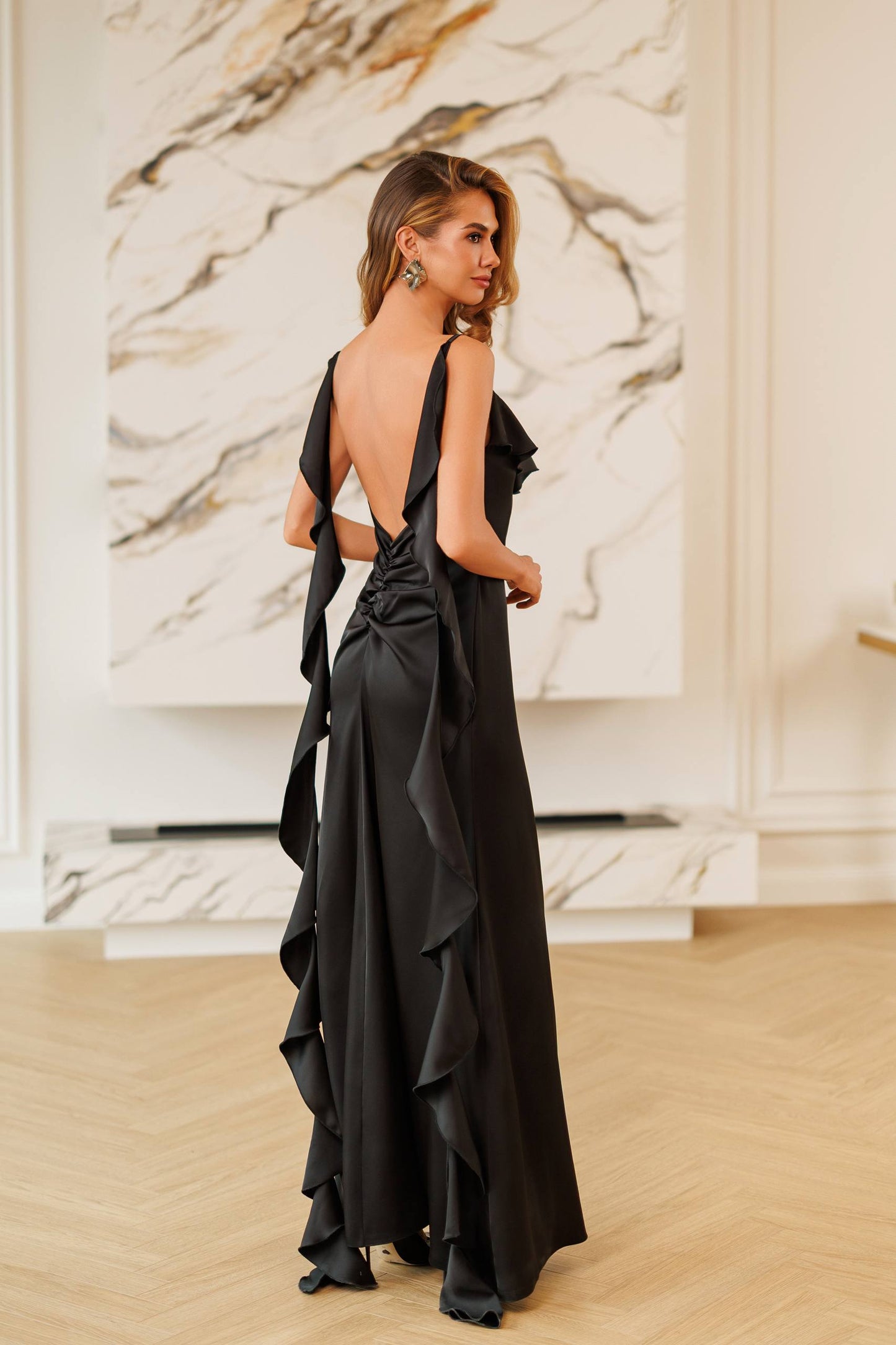 A woman wearing a black satin evening gown with a deep back and dramatic cascading ruffles, standing in a brightly lit room with a marble wall art piece and light wood floor.