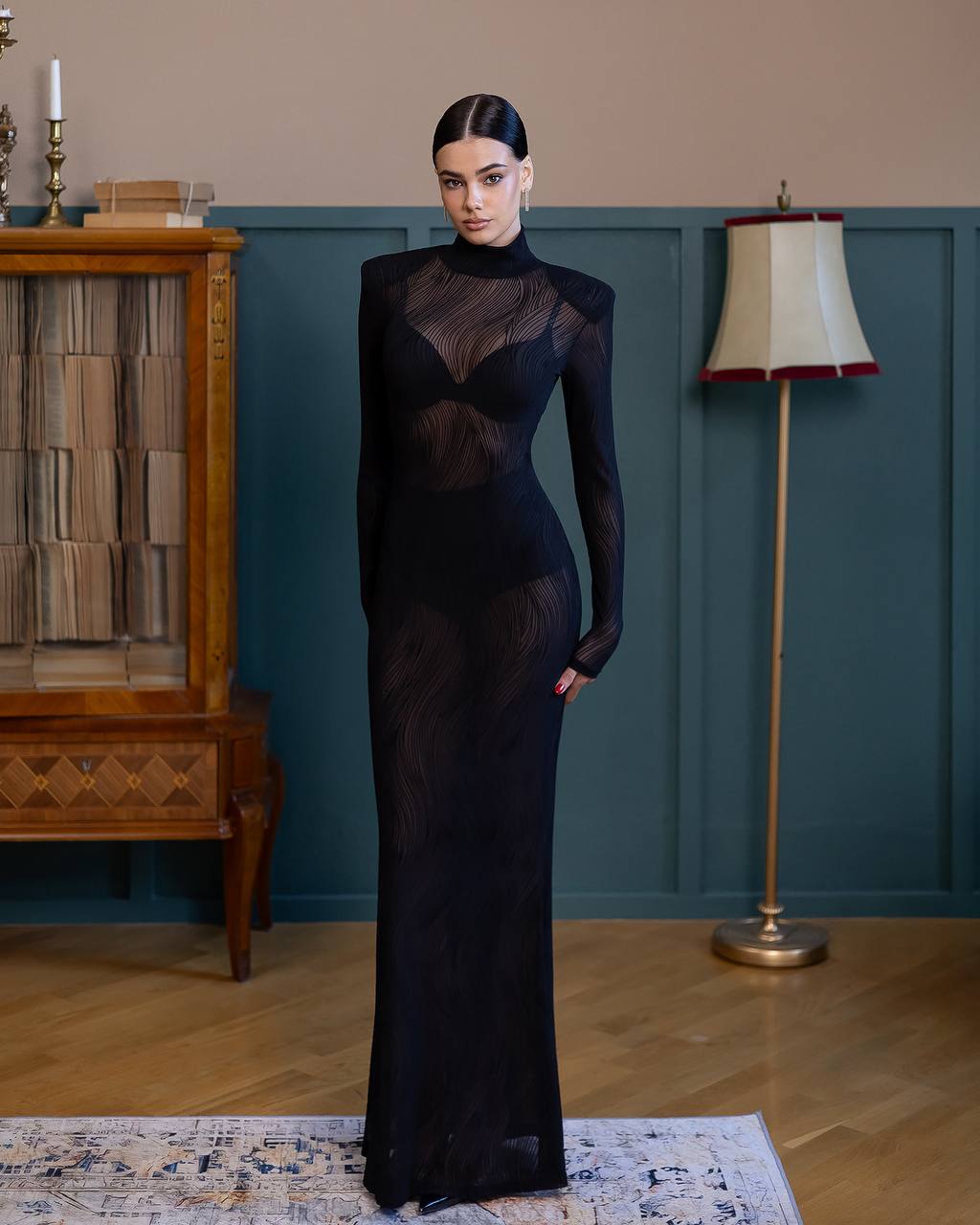 Woman in a black evening gown standing in a room with a lamp and wooden cabinet.