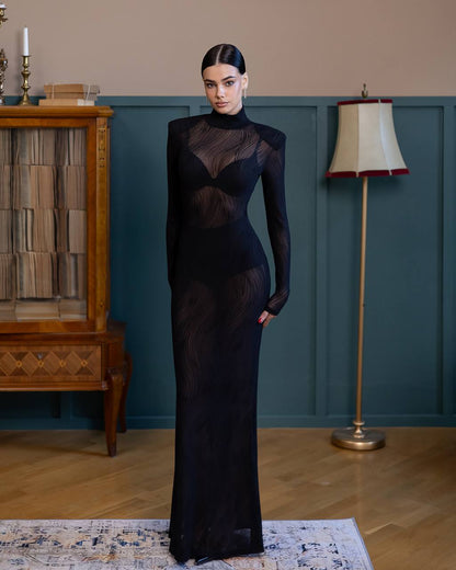 Woman in a black evening gown standing in a room with a lamp and wooden cabinet.