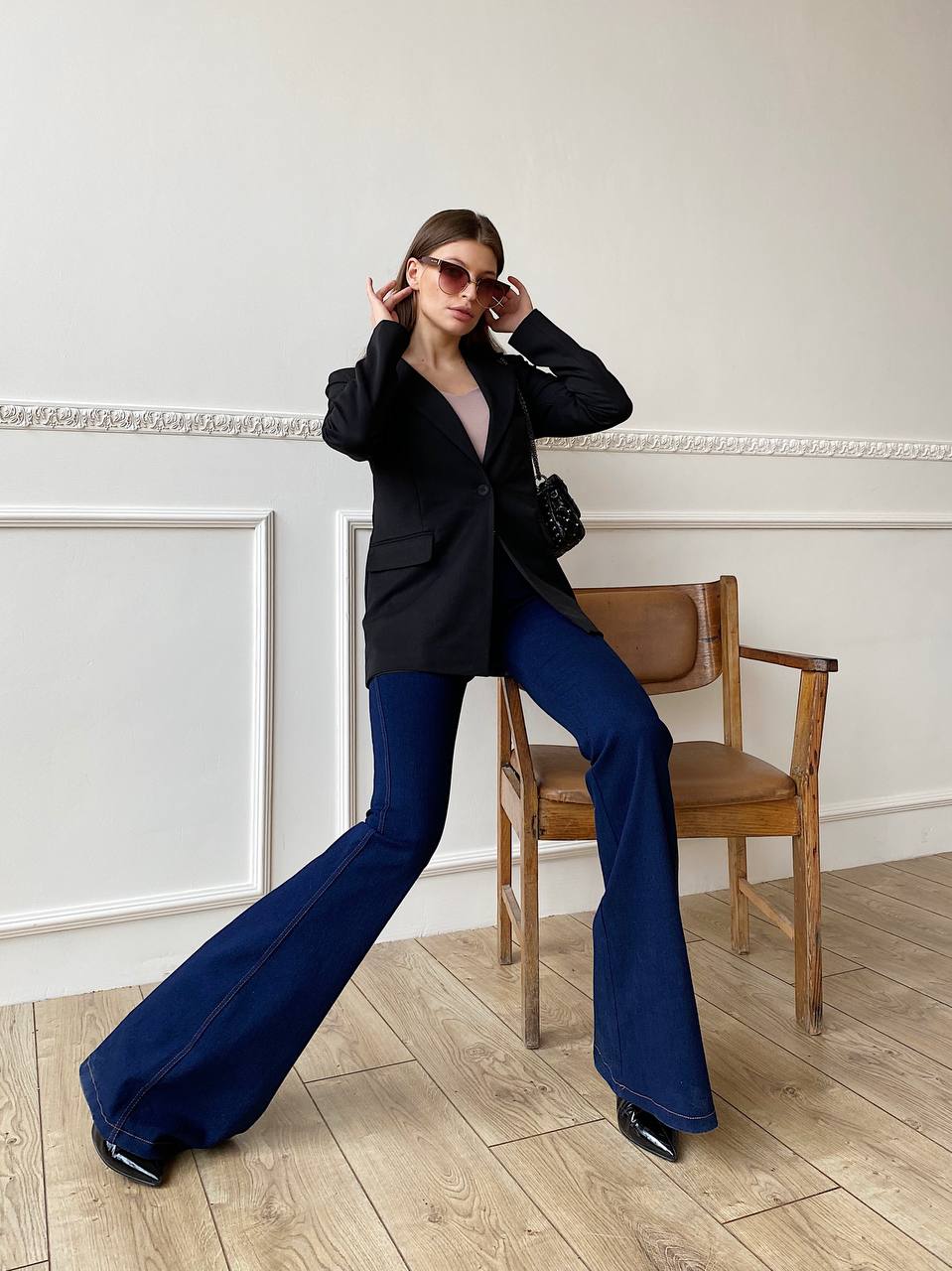 Woman in black blazer and blue pants posing in a room with wooden floor and white walls.
