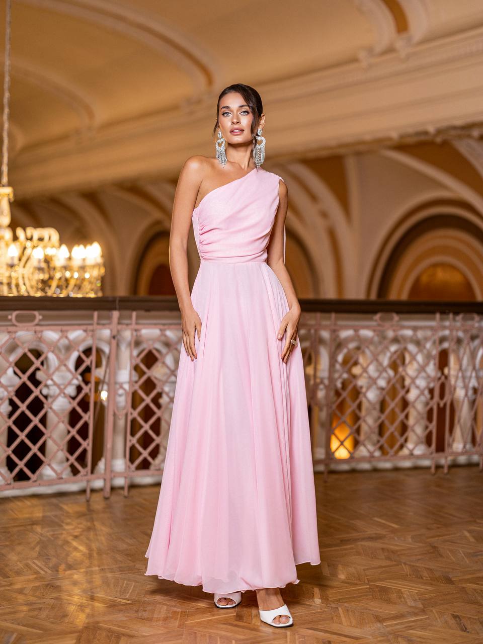 Woman in a pink one-shoulder dress standing in an elegant indoor setting with chandelier.