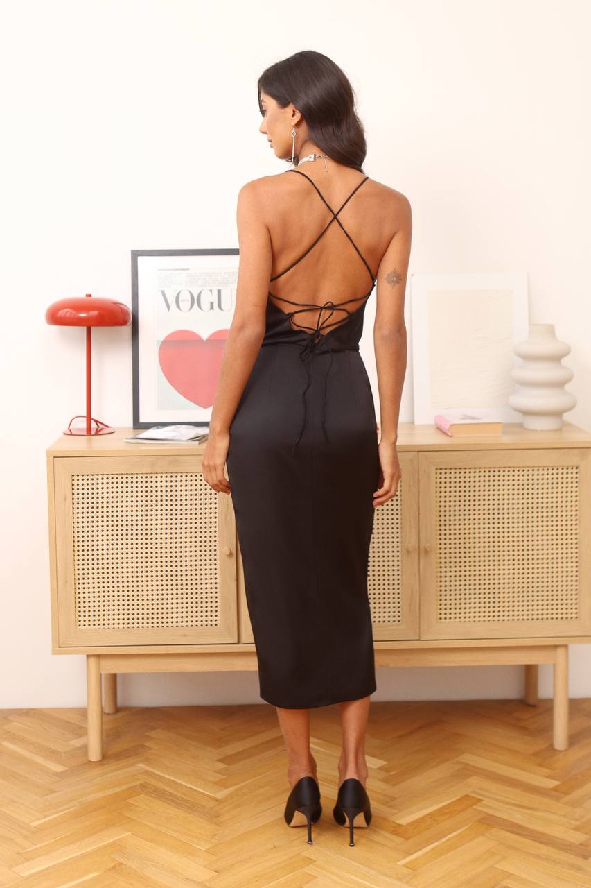 Woman wearing a black dress with criss-cross back design in a room with a wooden cabinet and decorative items.