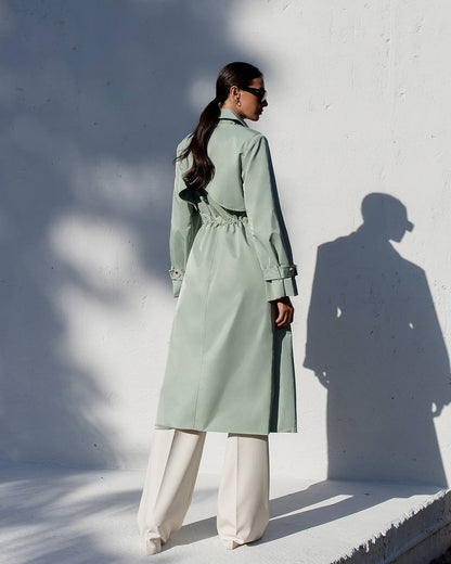 Person wearing a light green trench coat against a white wall with shadows.