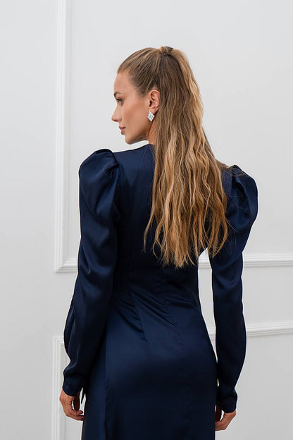 Woman wearing a navy blue dress with puffed sleeves against a white wall.