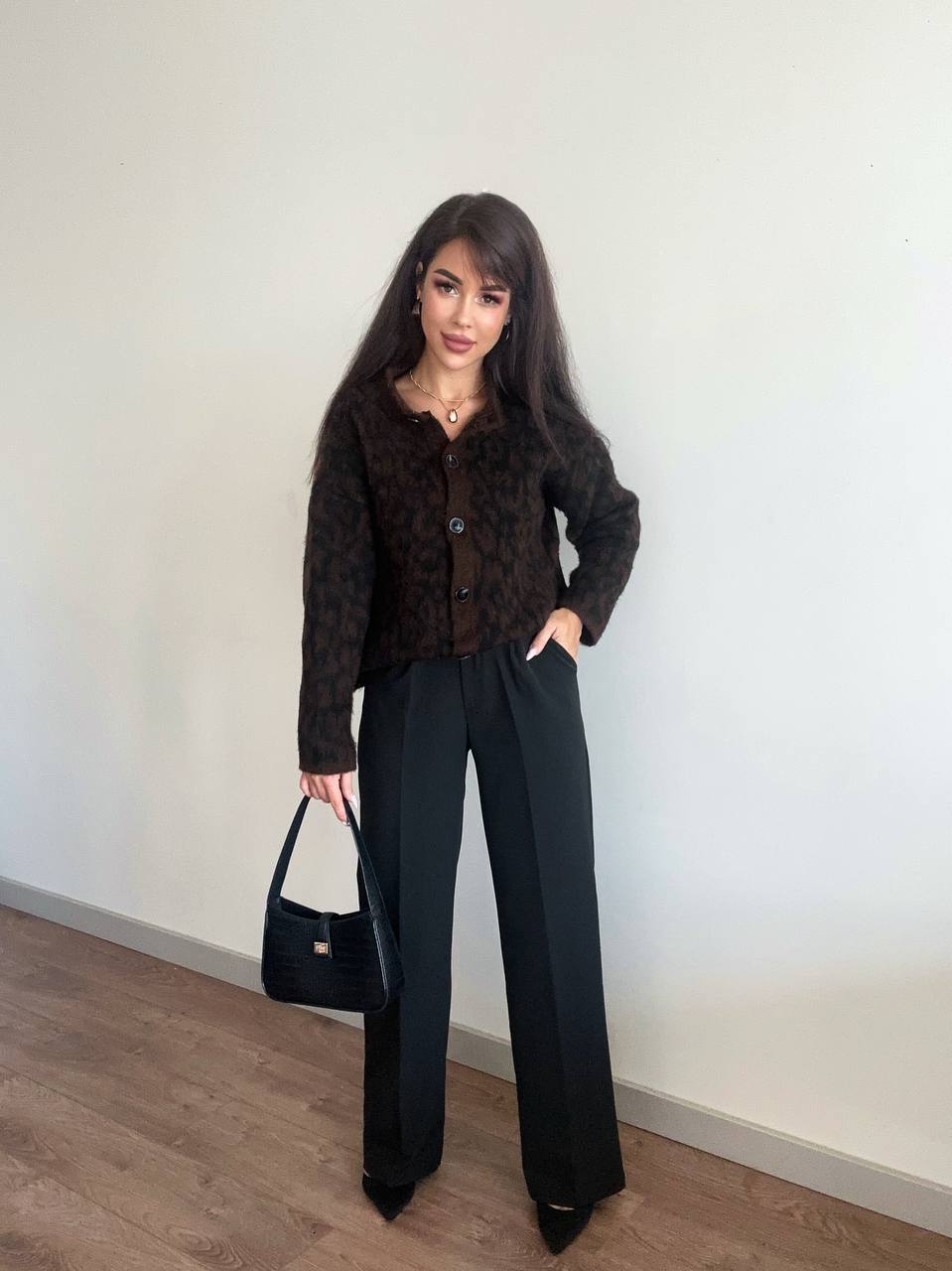 Woman wearing a brown patterned shirt and black pants, holding a black handbag against a white wall.