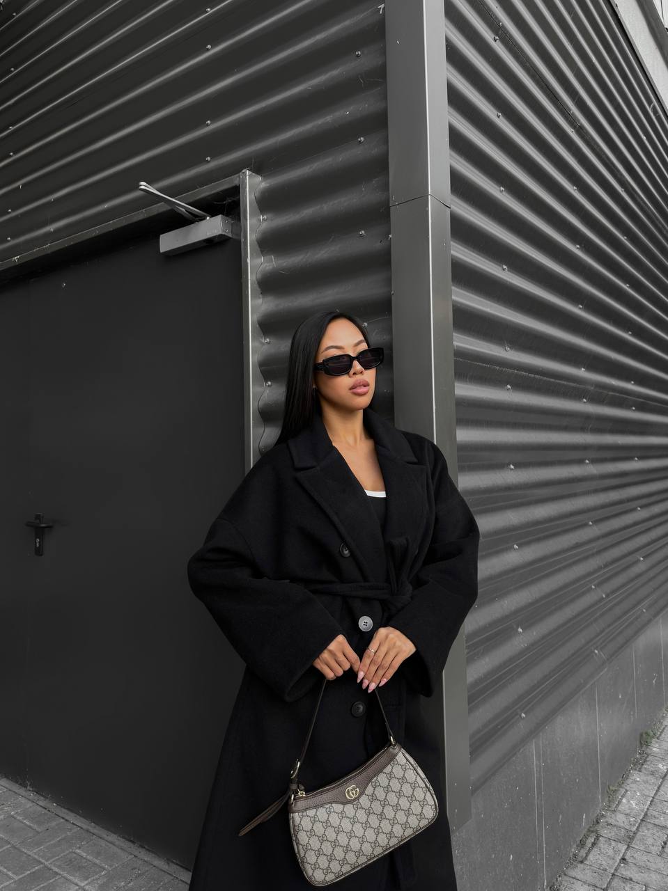 Woman in a black coat and sunglasses holding a handbag against a modern architectural background