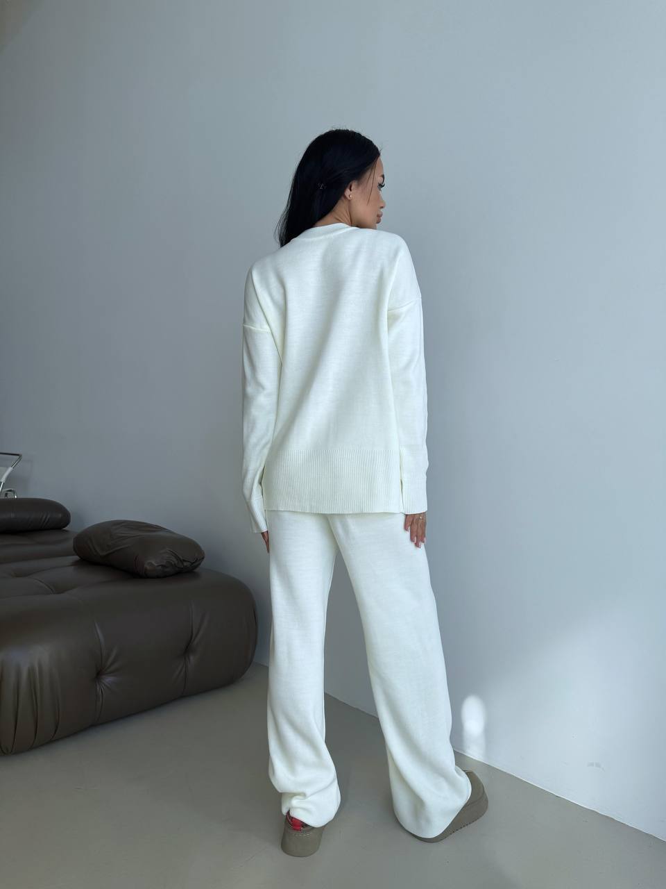 Person wearing a white outfit standing in a minimalistic room with a gray wall and brown sofa.