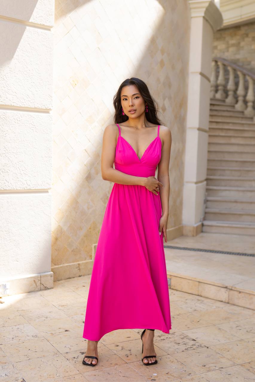 A woman wearing a bright hot pink maxi dress with spaghetti straps and a V-neckline, standing outdoors in front of a beige stone building with stairs.