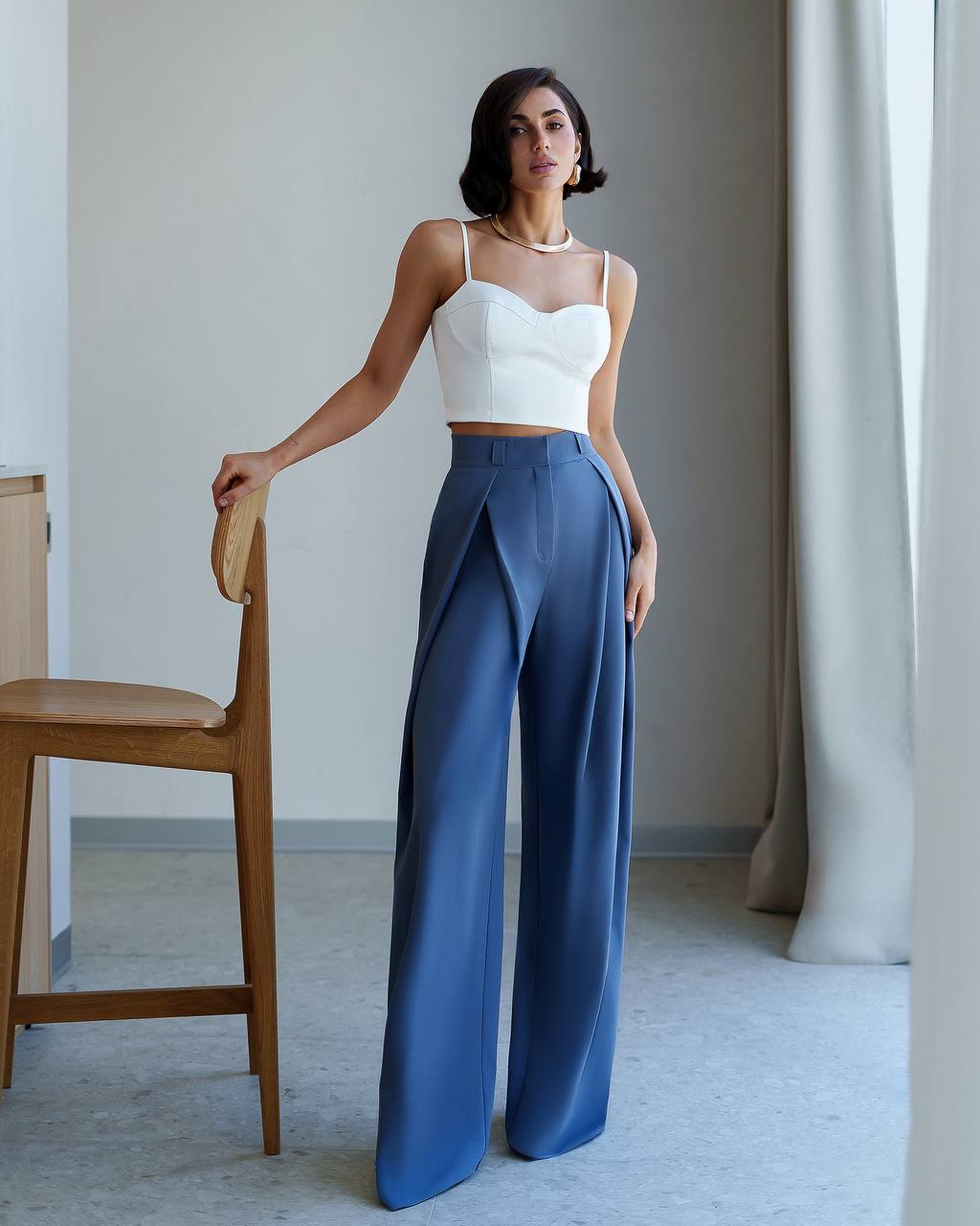Woman wearing a white top and blue pants standing in a room with a wooden chair and light-colored walls.