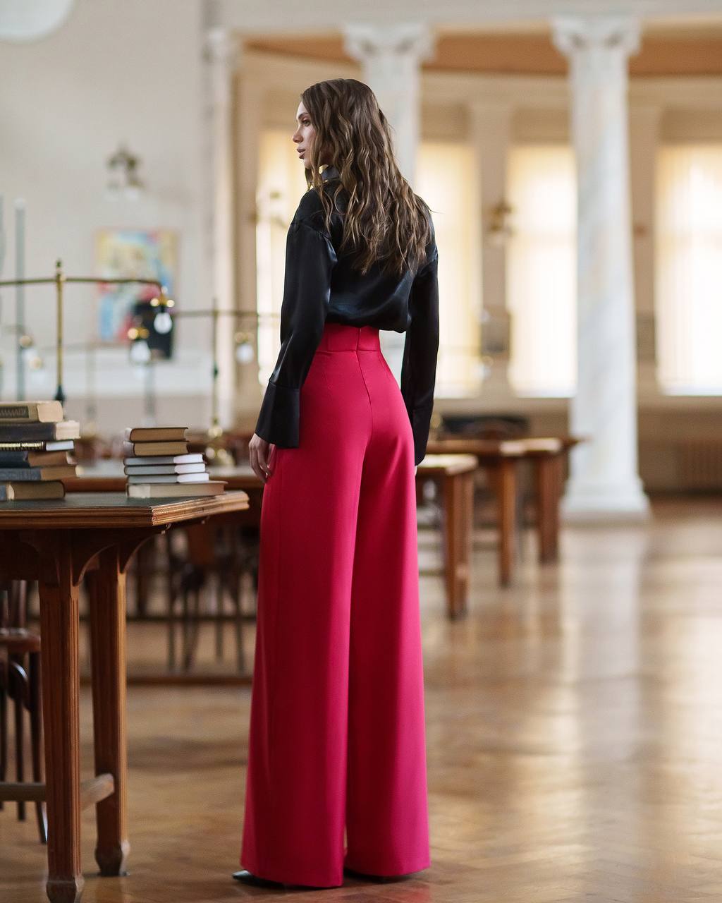 Woman wearing a black top and red pants standing in a library.