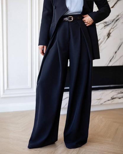 Person wearing a black coat and wide-leg pants in a room with marble wall.