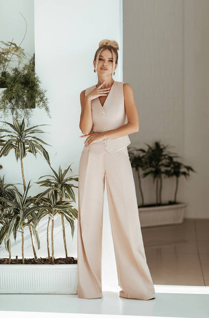 Woman in a beige sleeveless top and wide-leg pants standing in a room with plants.