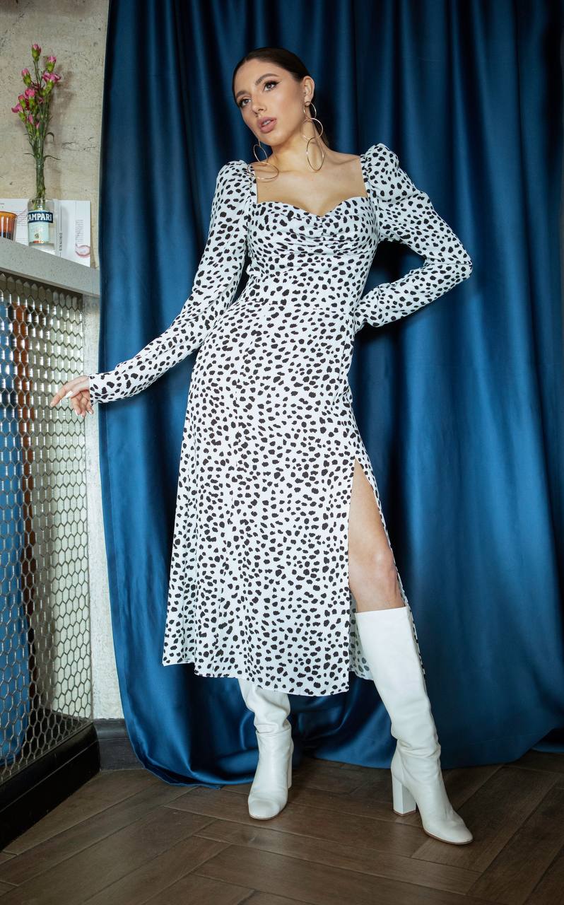 Woman wearing a black and white polka dot dress with a slit, standing against a blue curtain.