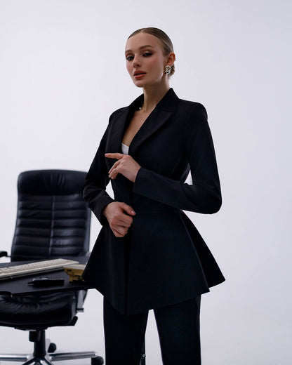 Woman in a black suit standing in an office setting with a chair and desk.