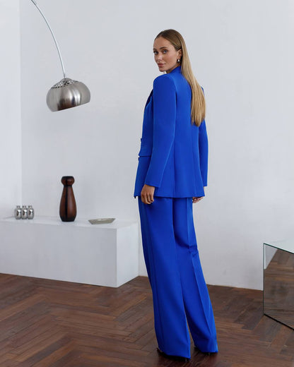 Woman in a blue suit standing in a modern interior setting with a white wall and wooden floor.
