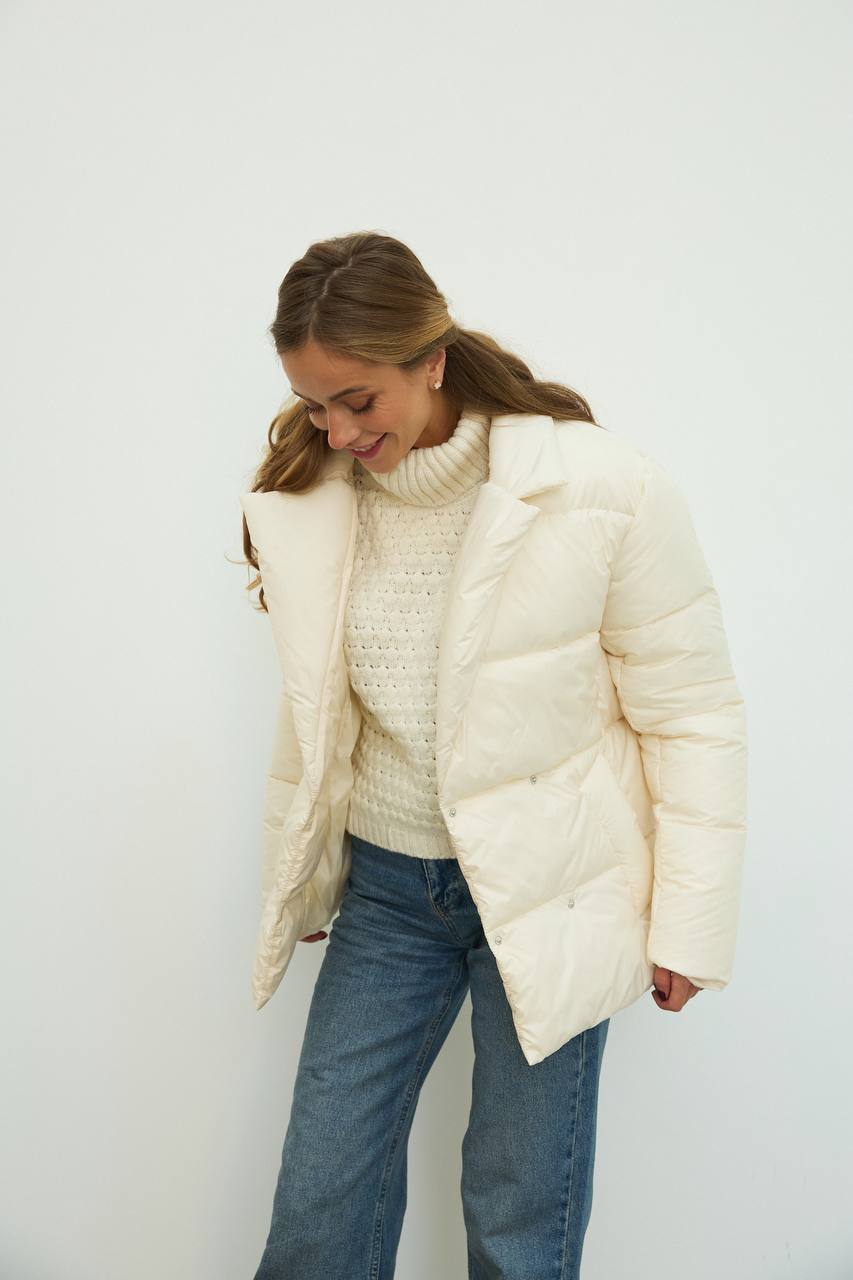 Woman wearing a white puffer jacket and sweater with blue jeans against a plain background