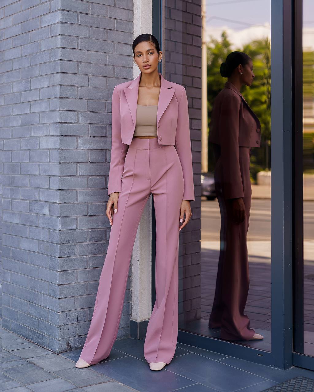 Woman in a pink suit standing against a brick wall with a mirror reflection.